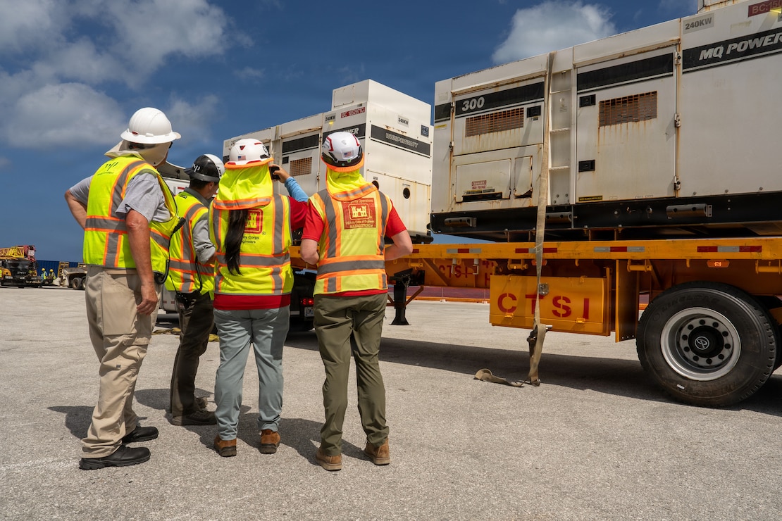 A group of individuals in hard hats and safety vests stand next to a flatbed truck loaded with large generators.