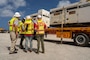 A group of individuals in hard hats and safety vests stand next to a flatbed truck loaded with large generators.