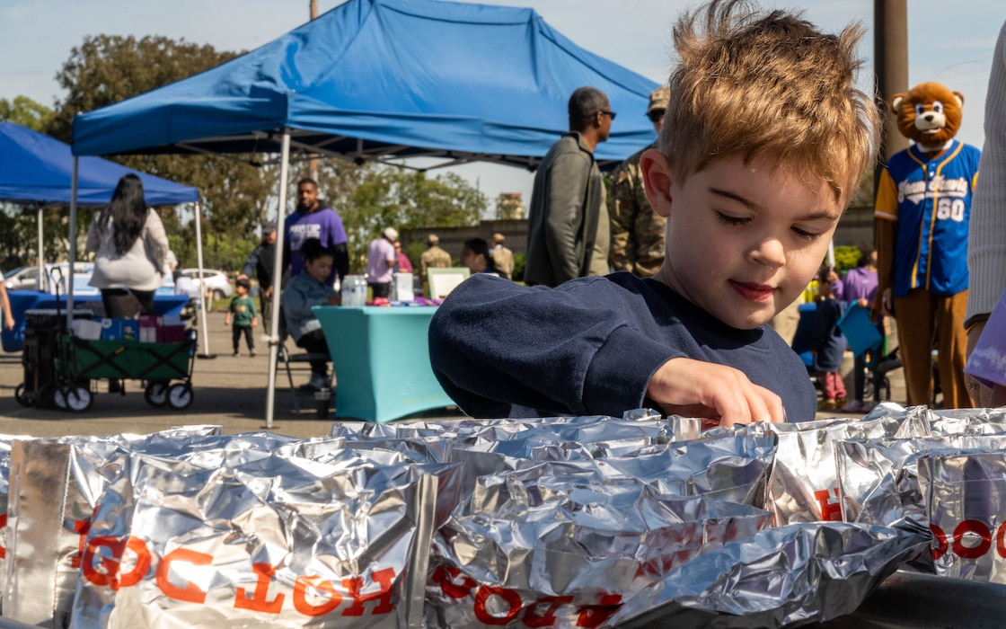 A boy selects a hot dog from a table with rows of hot dogs wrapped in silver foil, the word hot dog is written in red letters.