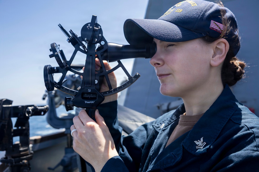 A sailor looks through a handheld instrument on the deck of a ship, with the horizon in the background.