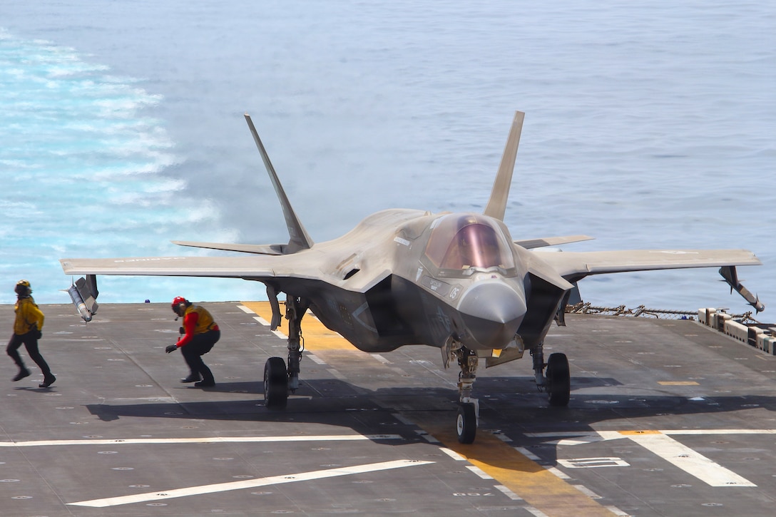 A fighter jet sits on the deck of a ship at sea, and two people stand near a wing.