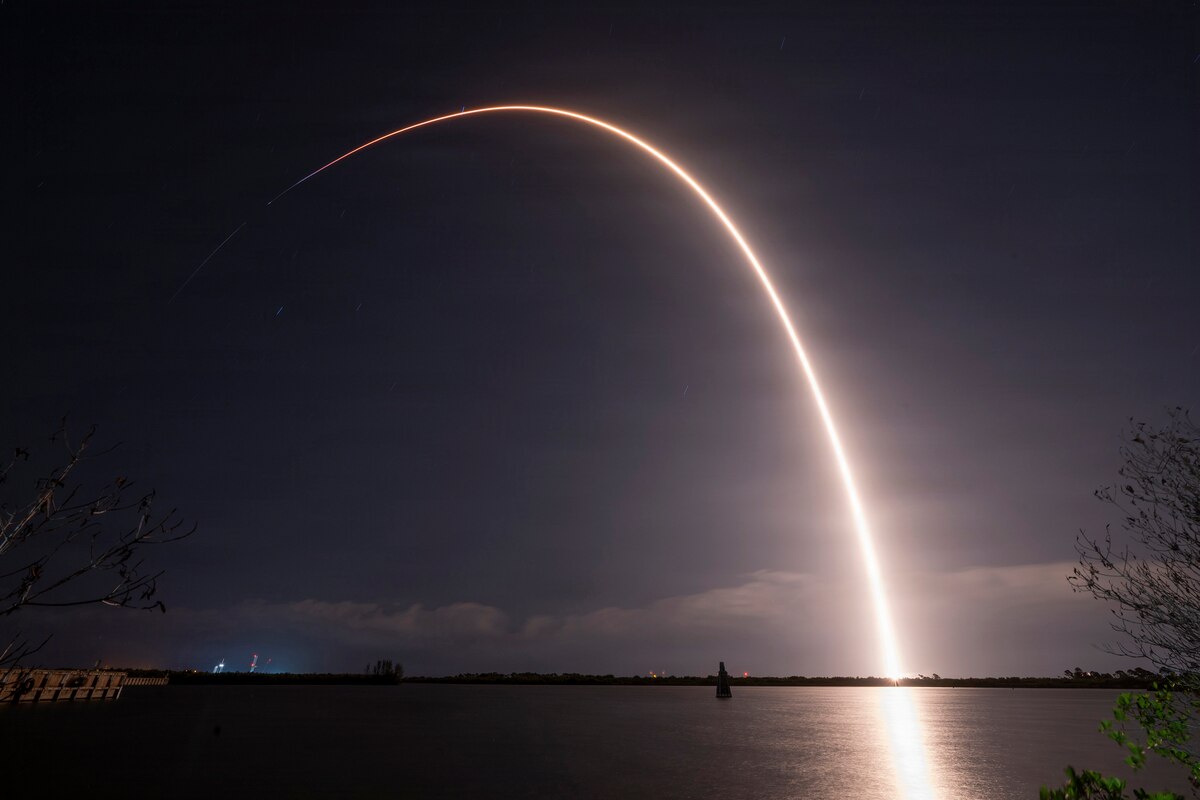 A rocket with a capsule on the end flies through the night sky, leaving a trail of white light reflecting off the water in the foreground.