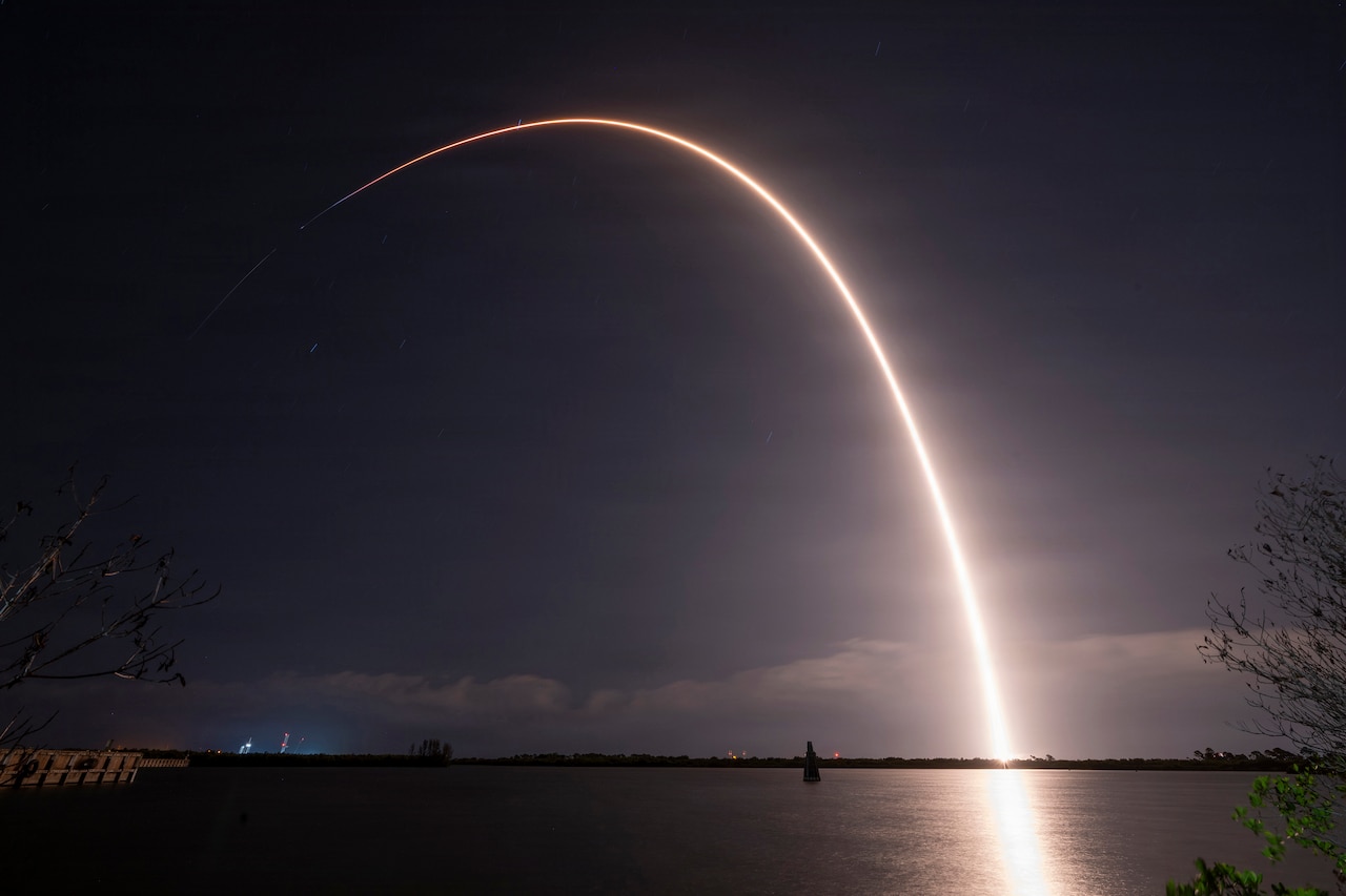 A rocket with a capsule on the end flies through the night sky, leaving a trail of white light reflecting off the water in the foreground.