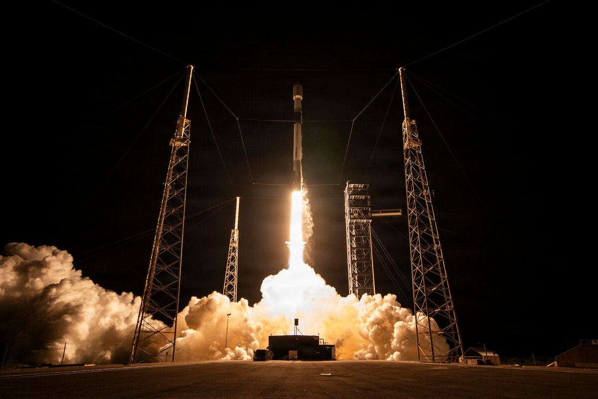 A rocket with a capsule on the end takes off into the night, shooting bright flames from the end and creating a cloud of smoke on the ground.