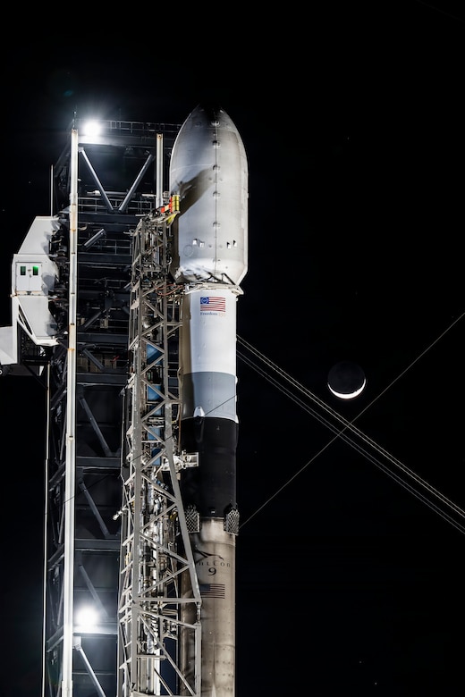 A rocket with a capsule on the end is positioned on the launching pad with the moon shown in the distant night sky.