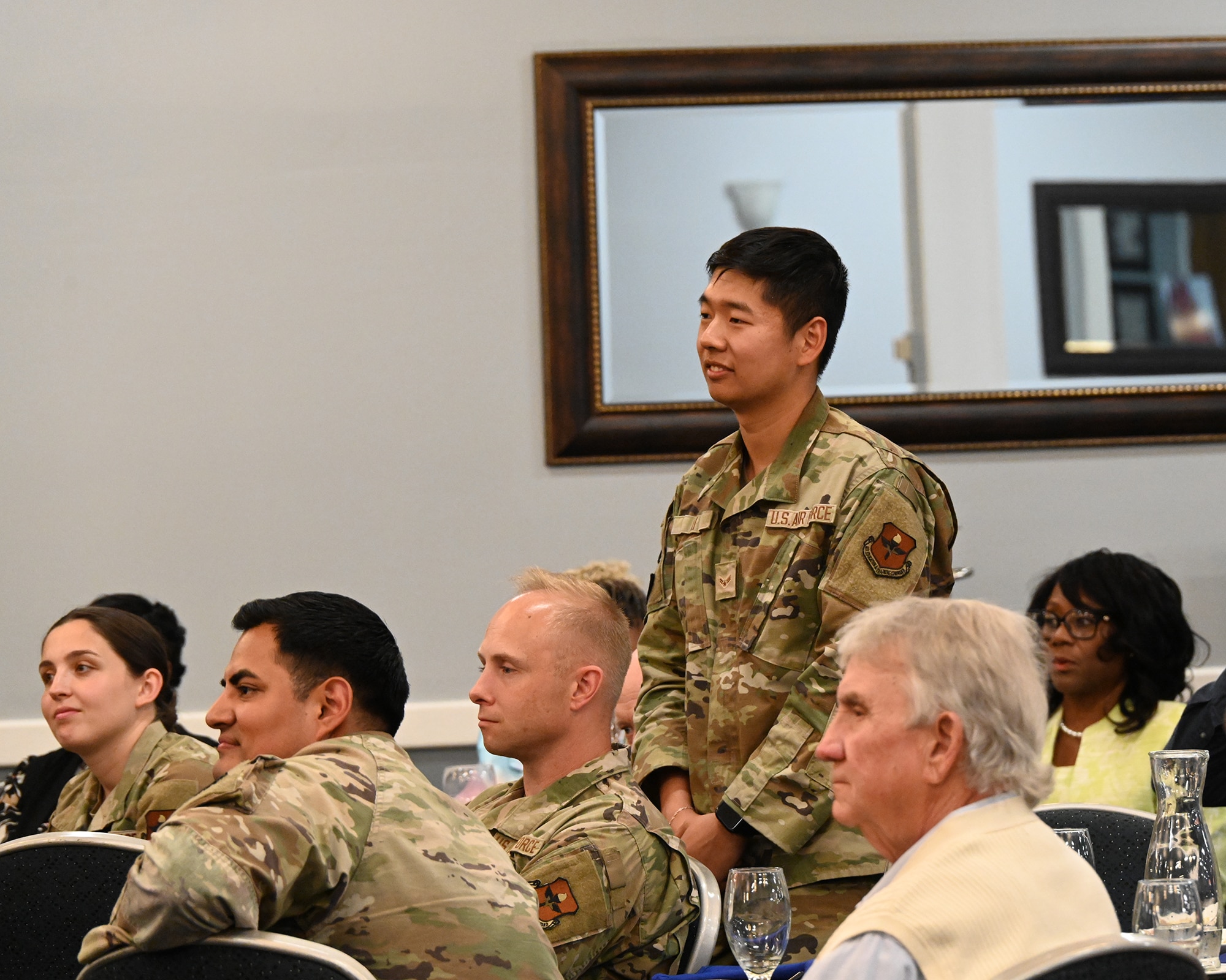 An Airman stands in a room full of people with a mirror behind him.