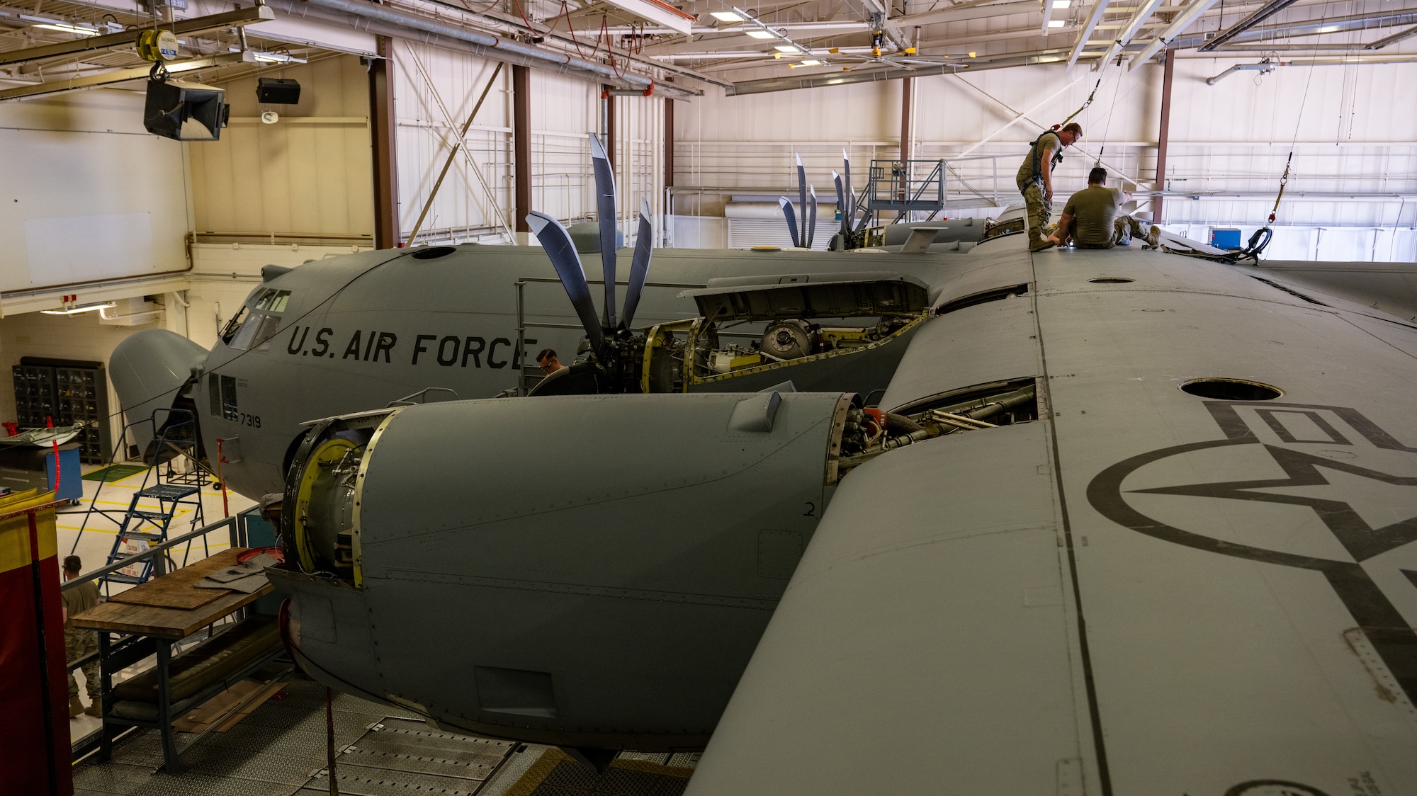 Airmen sit on top of a C-130H Hercules aircraft in safety harnesses performing an inspection.