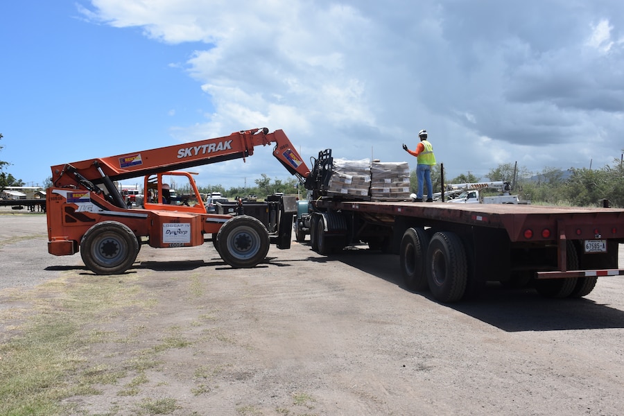 A man stands on the back of a flatbed truck as he directs a forklift operator who is lifting a pallet of supplies off the truck.