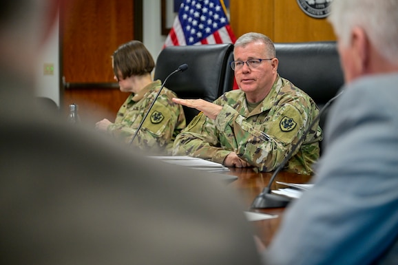 Two people in Army camouflage uniforms sit at the head of a conference table.