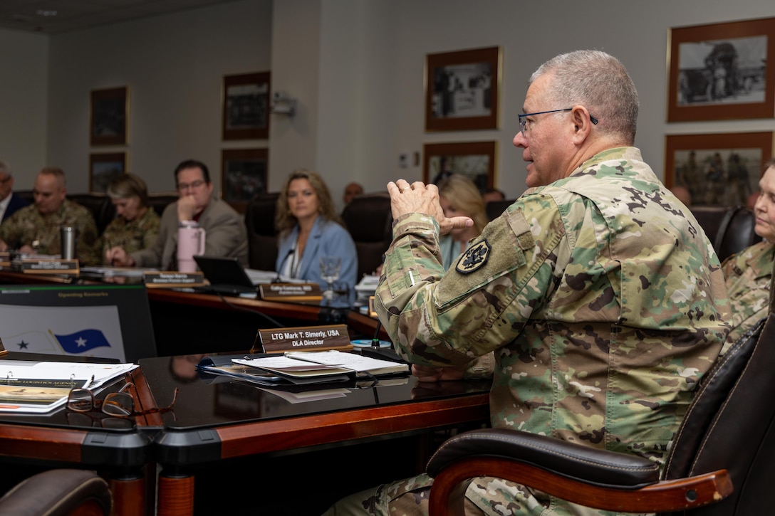 A man with graying hair and glasses in a camouflage military uniform speaks at a conference room table while others around the table listen.