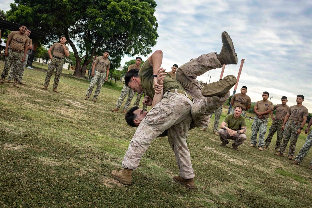 A Marine swings another Marine to the ground as dozens of Marines watch in a field under a blue sky.