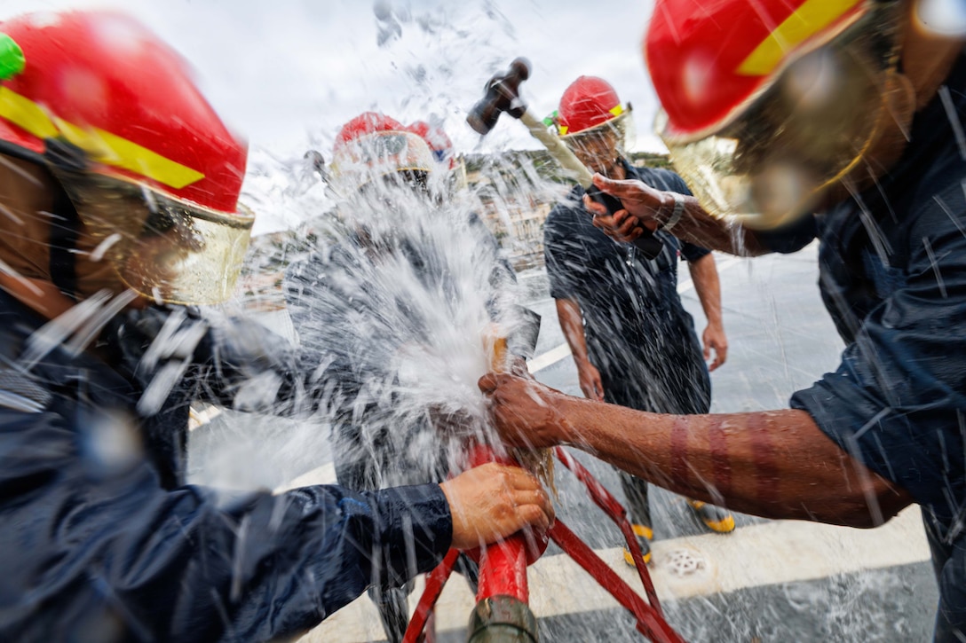 Sailors in blue jump suits and red helmets work on a ruptured pipe as water splashes everywhere.