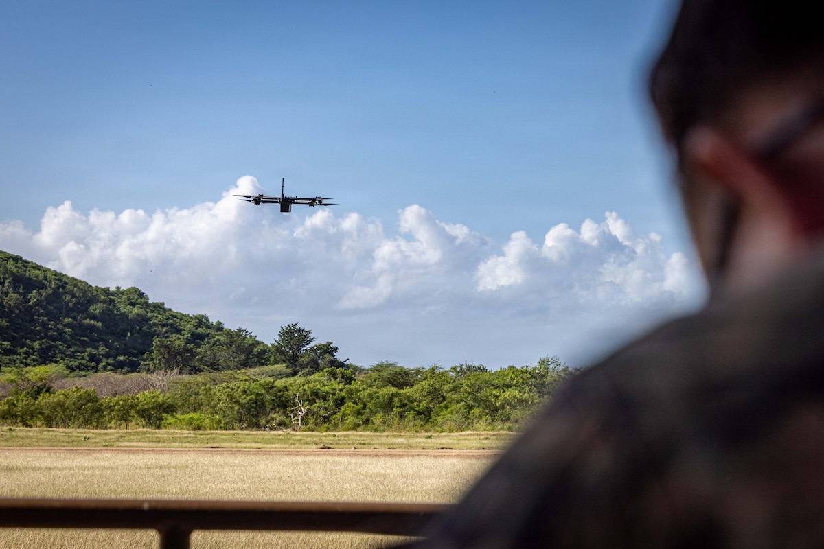 An unmanned aerial vehicle flies through a blue sky with clouds and trees below. In the foreground is the silhouette of a person with the controls.