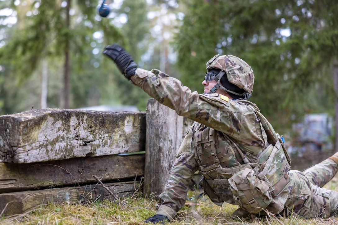 A soldier in tactical gear tosses a grenade over a wooden structure while lying on the ground in a wooded area.