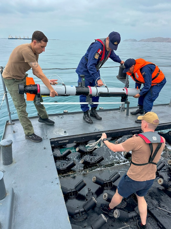 Two people in blue military uniforms and one person in casual attire stand on a boat and hold a long tube as they prepare to put it in the ocean; another person in casual attire is holding additional equipment and observing.