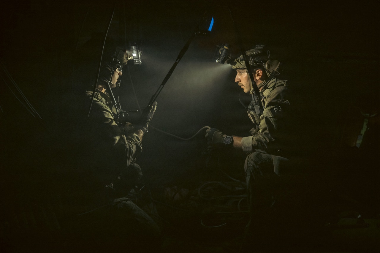 Two men in camouflage military uniforms and helmets with lights on the top look at each other while kneeling in a dark room; there is another person in a camouflage military uniform lying on the ground.