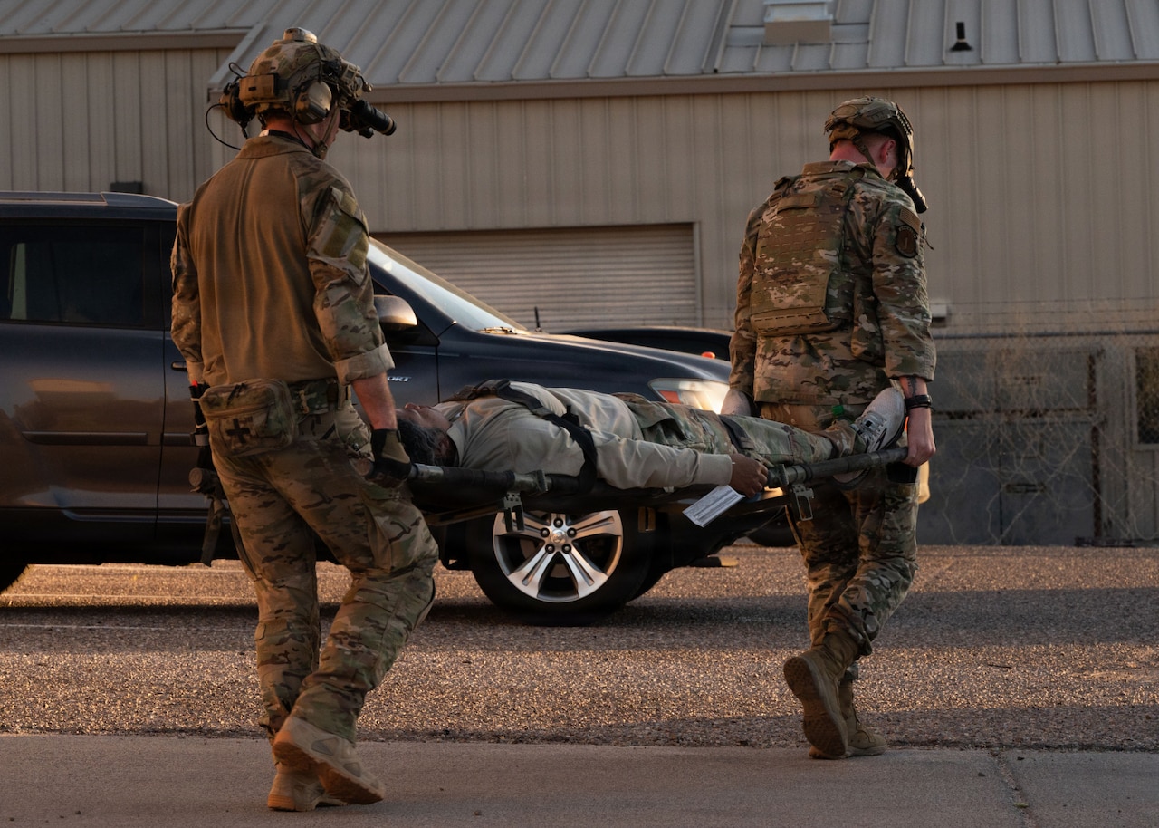 Two men in camouflage military uniforms carry a stretcher with another man in similar attire toward a building with a car parked in the background.