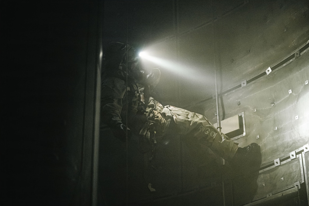 A person in a camouflage military uniform and a gas mask, rappels down a dark shaft.