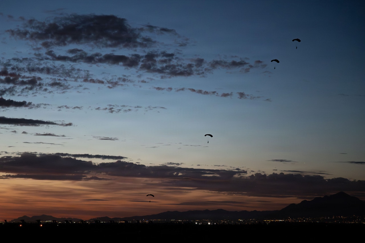 A wide shot of people gliding to the ground using parachutes as the sun sets; there are mountains in the background with lights from the surrounding area.