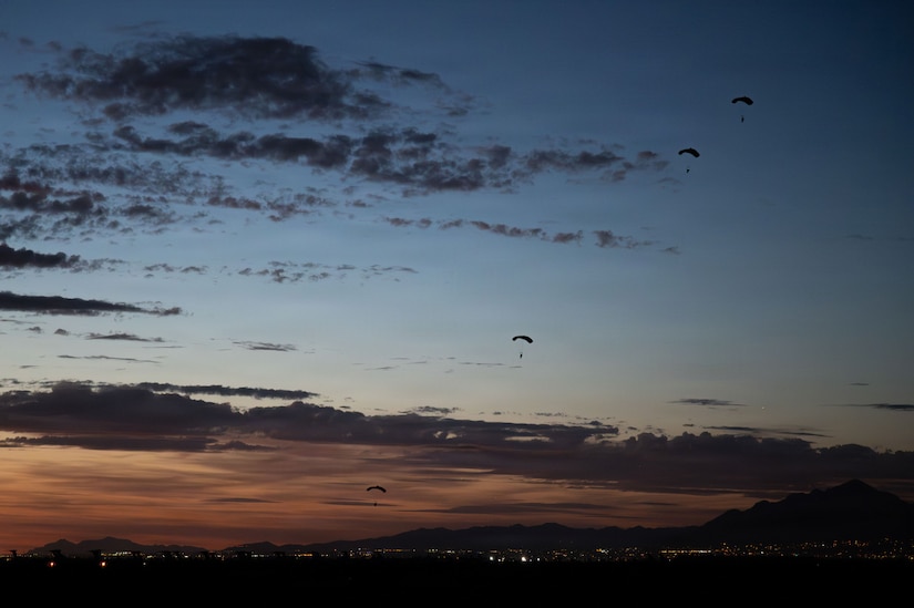 A wide shot of people gliding to the ground using parachutes as the sun sets; there are mountains in the background with lights from the surrounding area.