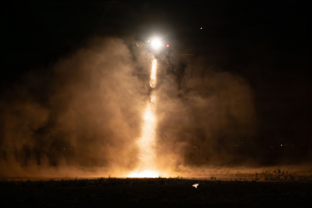 A military helicopter hoovers while shining a light on the ground, illuminating the dust in the air.