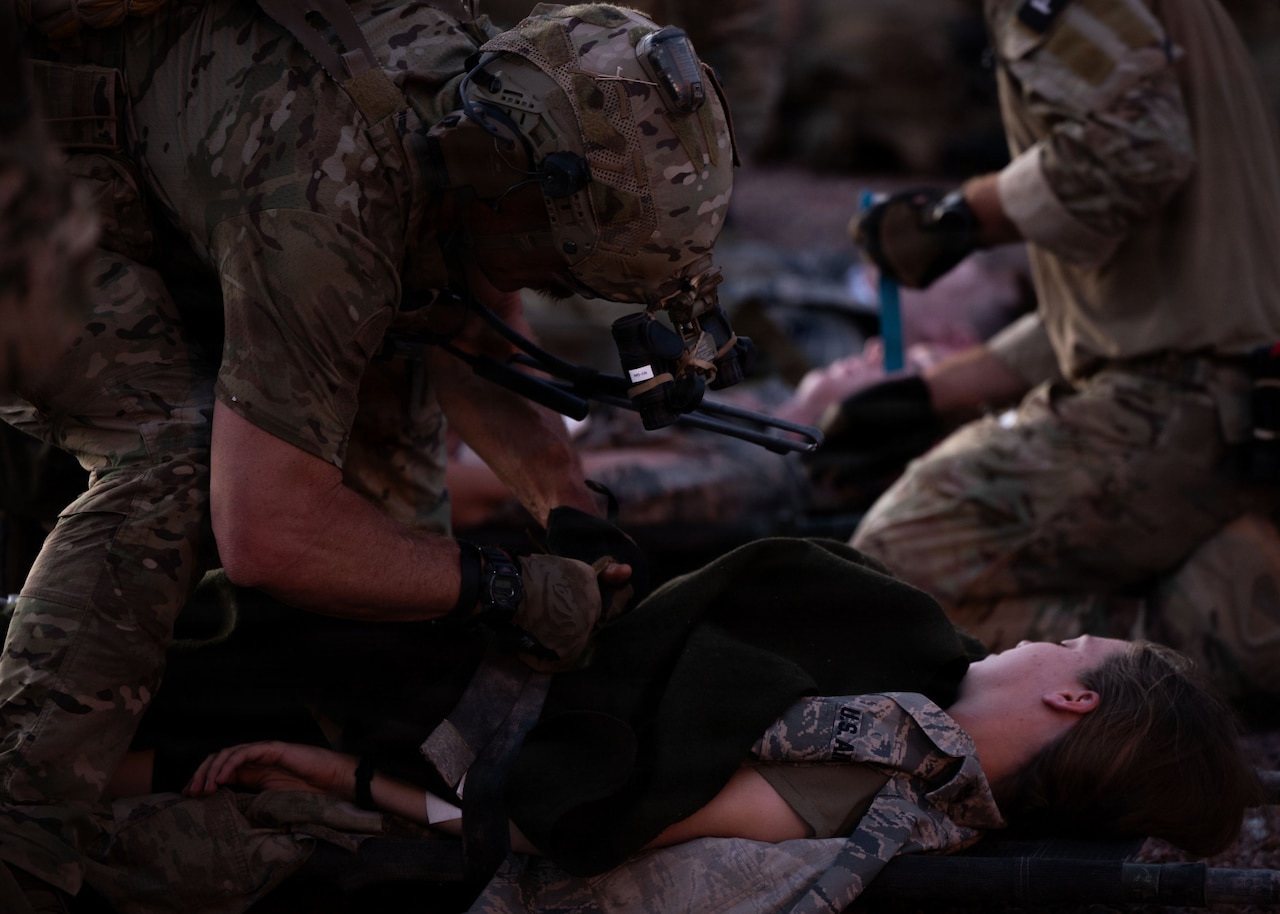 A man in a camouflage military uniform and helmet treats the simulated wounds of a woman in similar attire lying on the ground; people in similar attire are doing the same thing in the background.