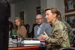 A woman with short red hair in a Army camouflage uniform speaks in a meeting.