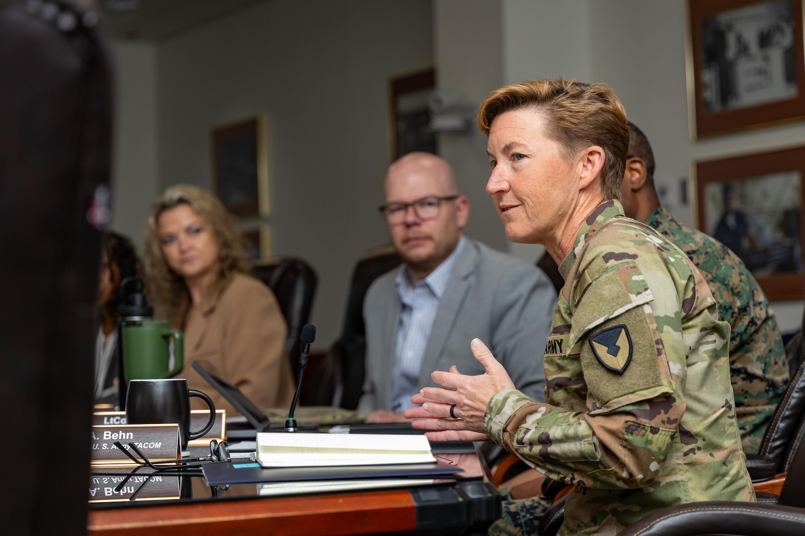 A woman with short red hair in a Army camouflage uniform speaks in a meeting.
