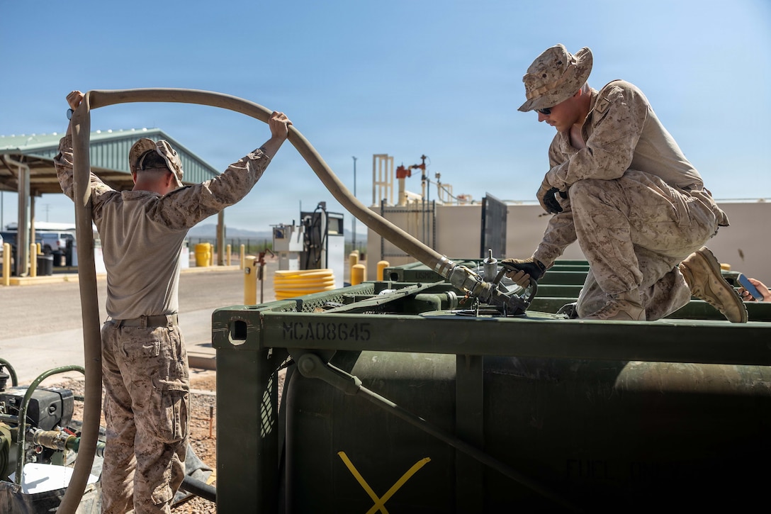 One service member holds up a fuel line while another service member holds the nozzle attached to it while kneeling on top of a large container.