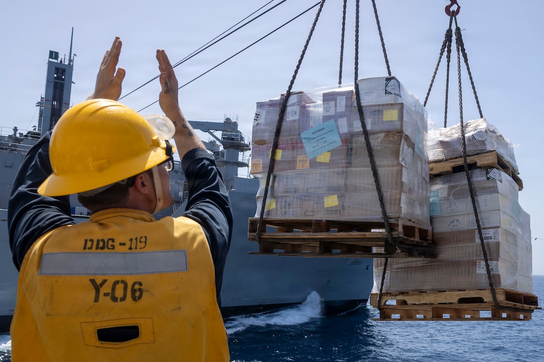A sailor facing cargo attached to cables off the side of a military ship raises their arms to give a signal.