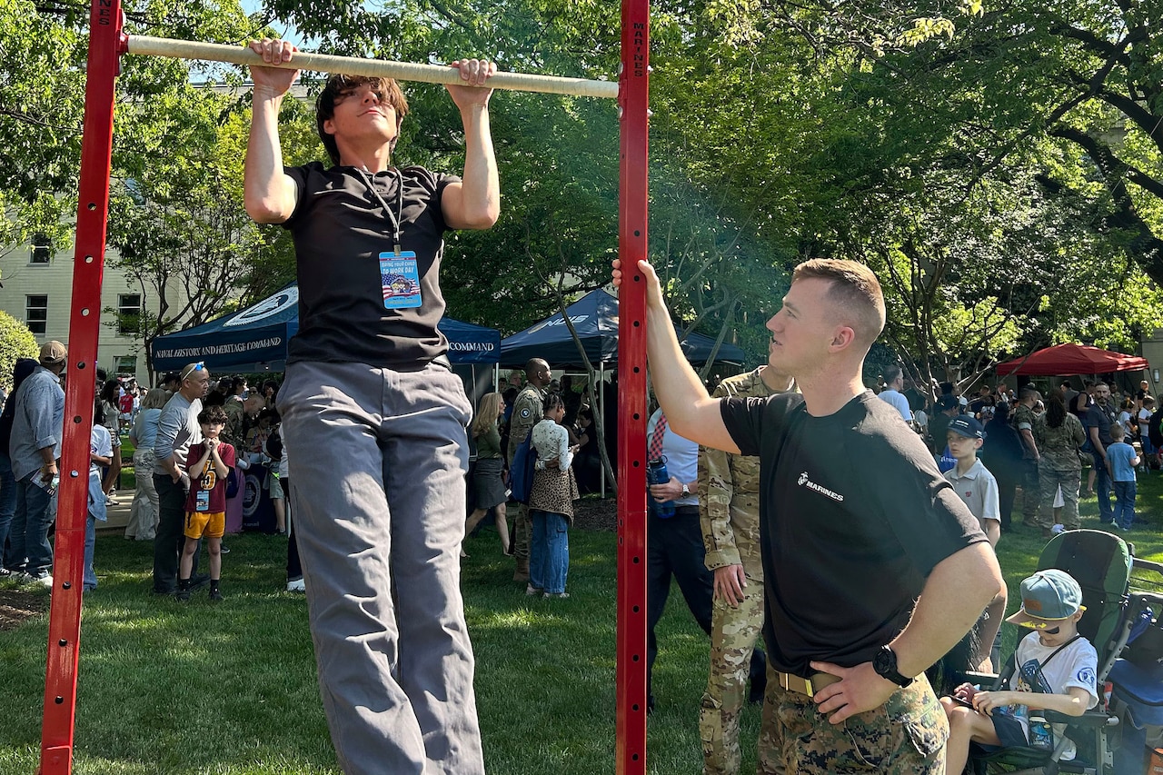 A young man holds onto a metal bar while pulling himself up. A man wearing camouflage military pants watches him. A crowd of people are in the background.