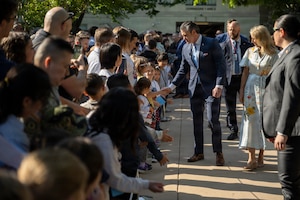 A man in a navy-blue suit shakes hands with a child in a crowd. A woman wearing a spring floral dress walks beside him.