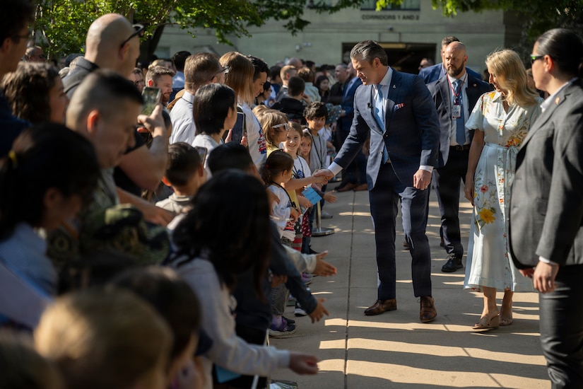A man in a navy-blue suit shakes hands with a child in a crowd. A woman wearing a spring floral dress walks beside him.