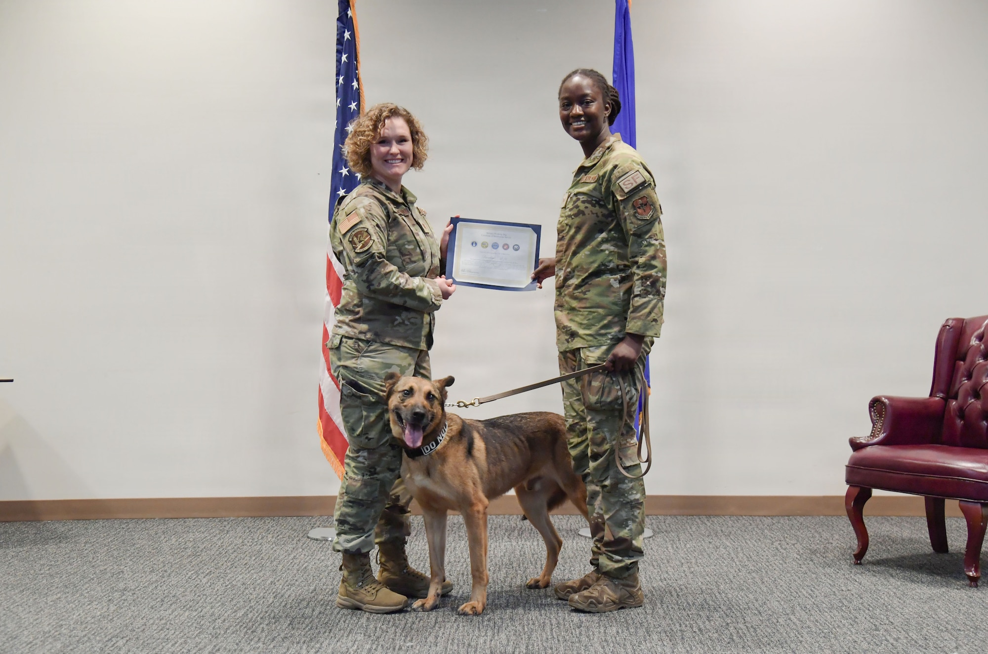 Two women in military uniform hold a certificate with a dog standing near them.