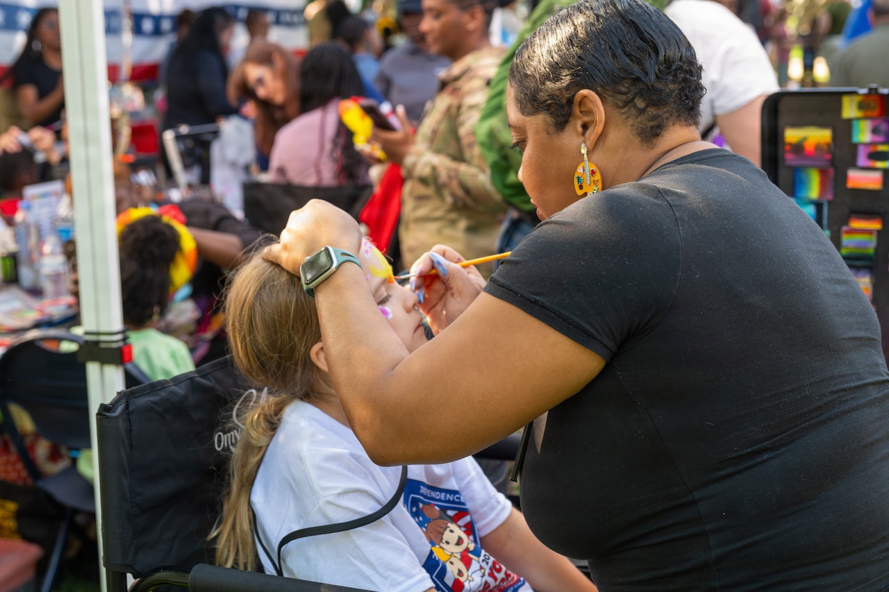 A woman with a short, cropped haircut, wearing a black shirt, sits while she paints flowers onto a young girl's face.