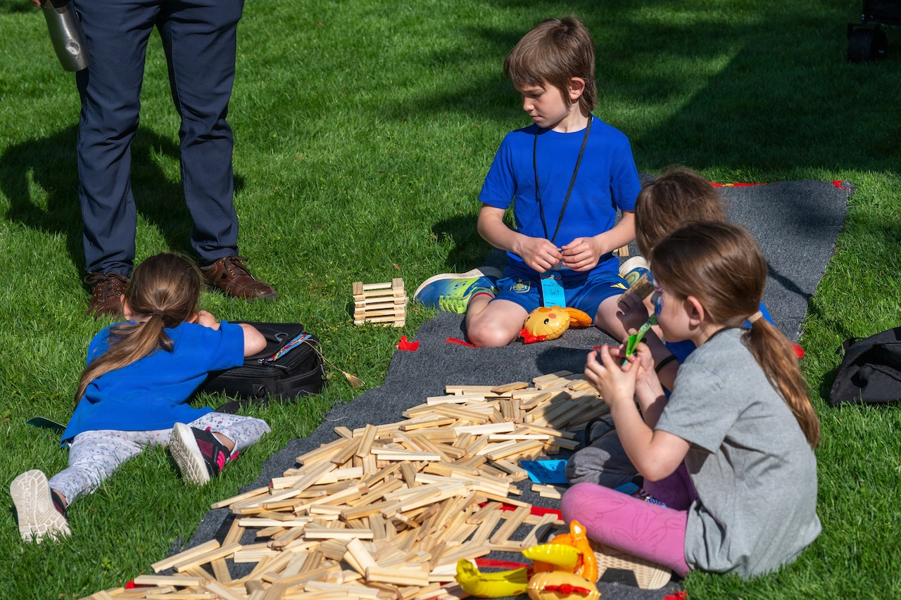 Four children sit on a lawn and play with wooden blocks. An adult stands nearby.
