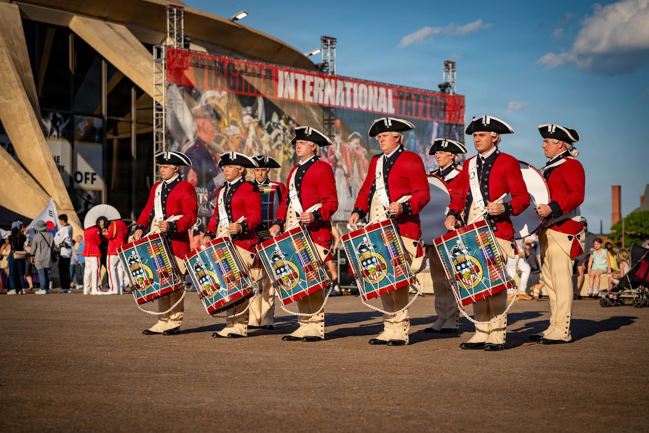 Troops in red historical uniforms beat ceremonial drums as they march in a parade through a city.