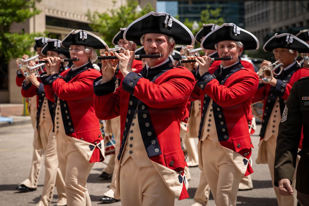 Troops in red historical uniforms play various musical instruments as they march in a parade through a city.