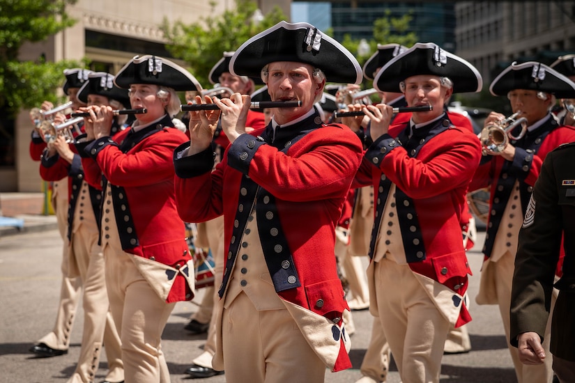 Troops in red historical uniforms play various musical instruments as they march in a parade through a city.