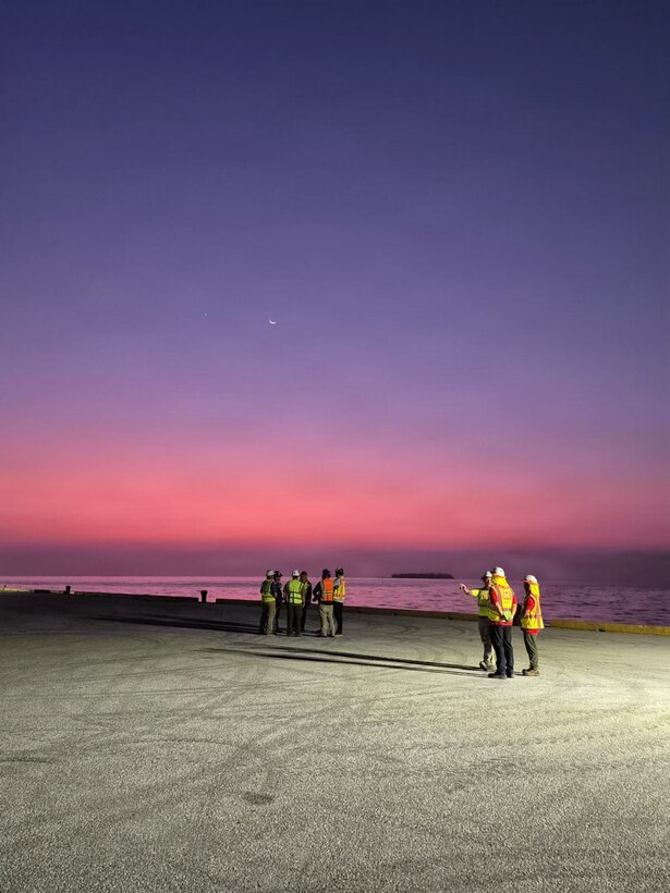 the sun is beginning to rise as people are seen standing near the water on a pier where ships are offloaded or loaded.