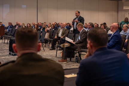 A man in business attire stands and speaks into a microphone. All around him, people are seated in an audience.