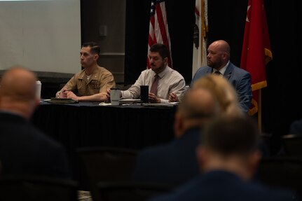 Two men in business attire sit at a table with a man in a formal military uniform as they listen to questions from the audience seated in front of them; behind them are an American flag and two other flags.