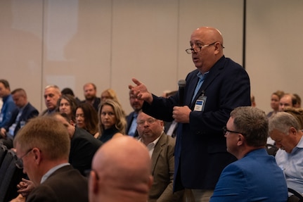 A man in business attire stands and speaks into a microphone. All around him, people are seated in the audience.
