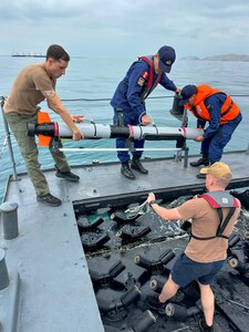 Two people in blue military uniforms and one person in casual attire stand on a boat and hold a long tube as they prepare to put it in the ocean; another person in casual attire is holding additional equipment and observing.
