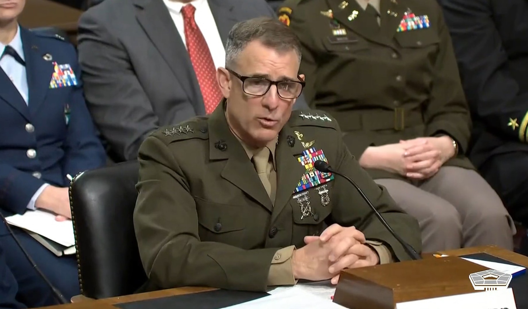 A man in a formal military uniform sits at a desk and speaks into a microphone. People in similar attire are behind him.
