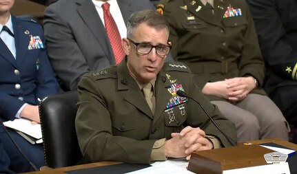 A man in a formal military uniform sits at a desk and speaks into a microphone. People in similar attire are behind him.