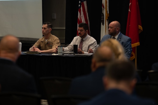 Two men in business attire sit at a table with a man in a formal military uniform as they listen to questions from the audience seated in front of them; behind them are an American flag and two other flags.