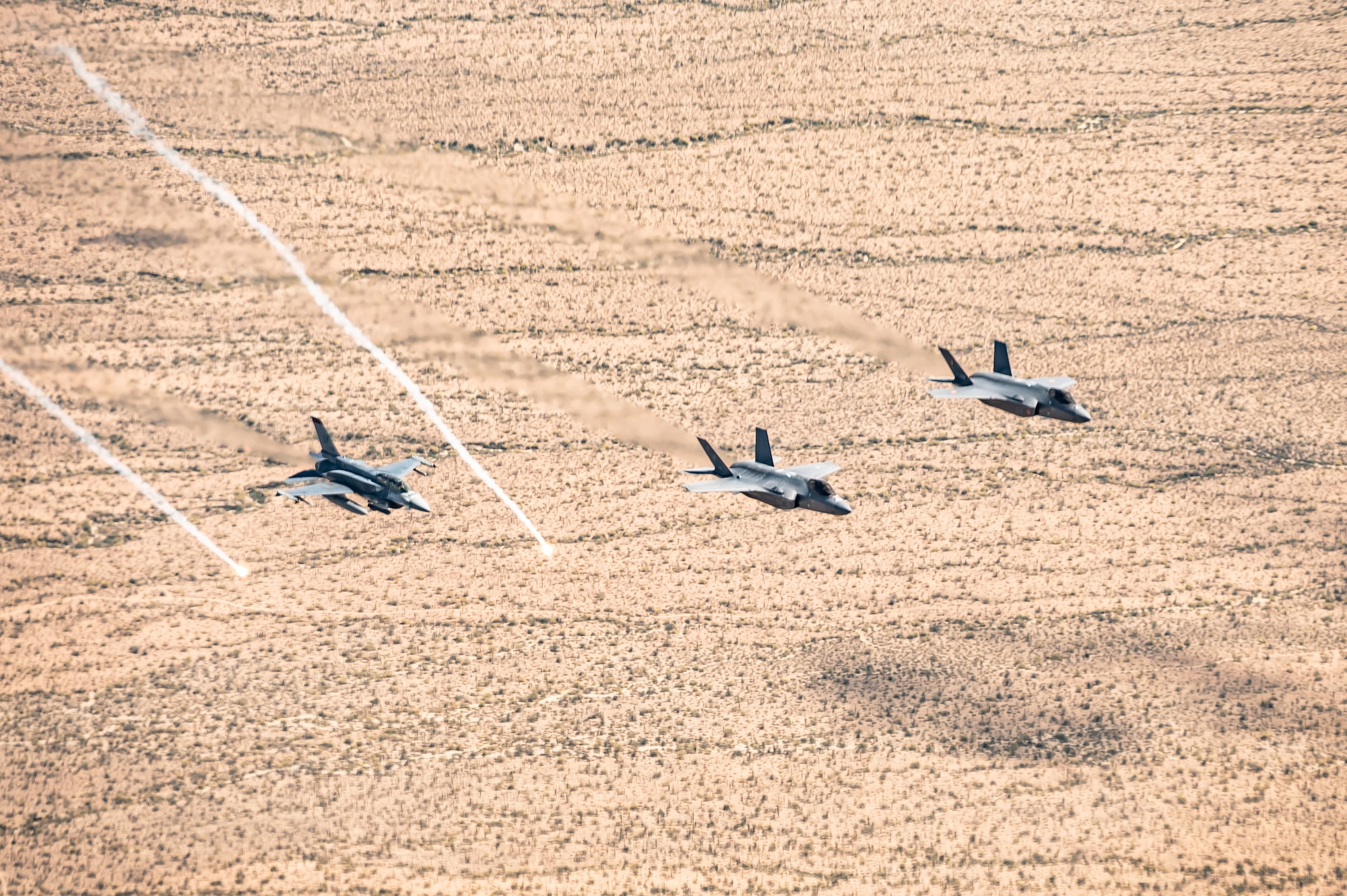 From left, a Republic of Singapore Air Force F-16D Fighting Falcon, U.S. Air Force F-35A Lightning II, and Belgian Air Force F-35A Lightning II, fly in formation as flares are released over the Sonoran Desert