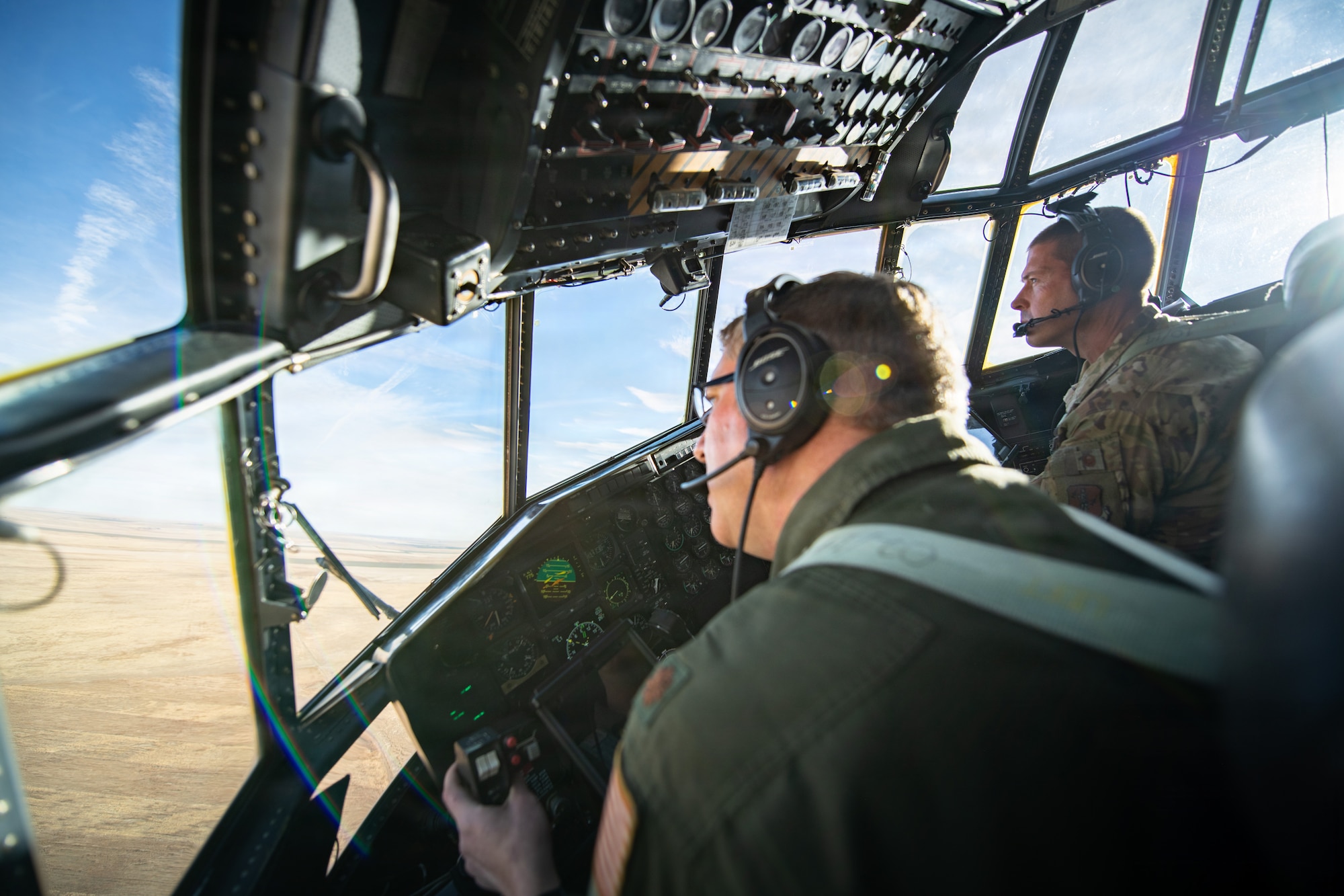 The sun shines through as a pilot looks out from a panel of windows from the flight deck of a C-130 Hercules onto the sky and ground below.