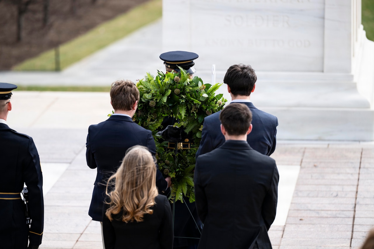 The back of four young adults in business attire is shown as they pass a wreath to a man in a military dress uniform. Another man in a military dress uniform stands off to the left.